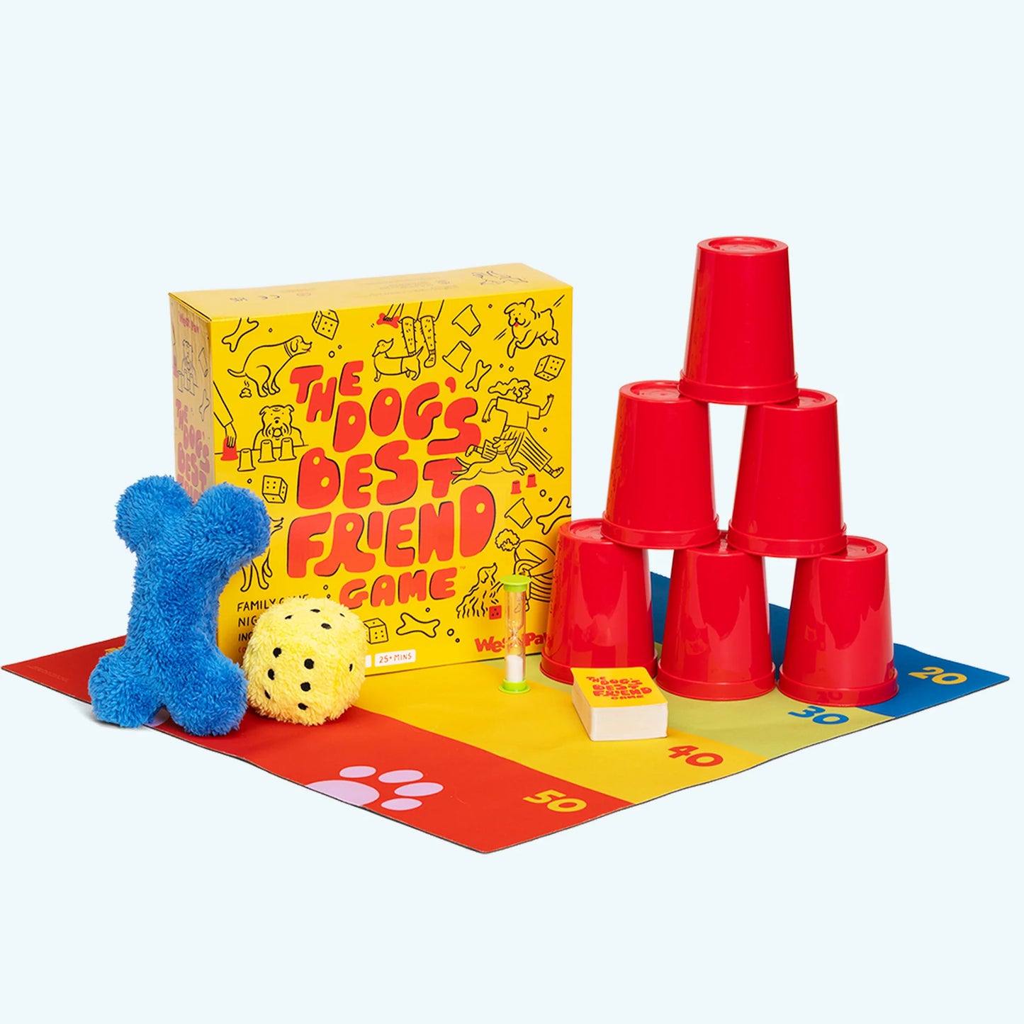 "The Dog's Best Friend" board game box with a blue plush bone toy, a yellow plush dice toy, 6 red cups stacked in a tower shape, a sand timer, playing cards, all on a play mat