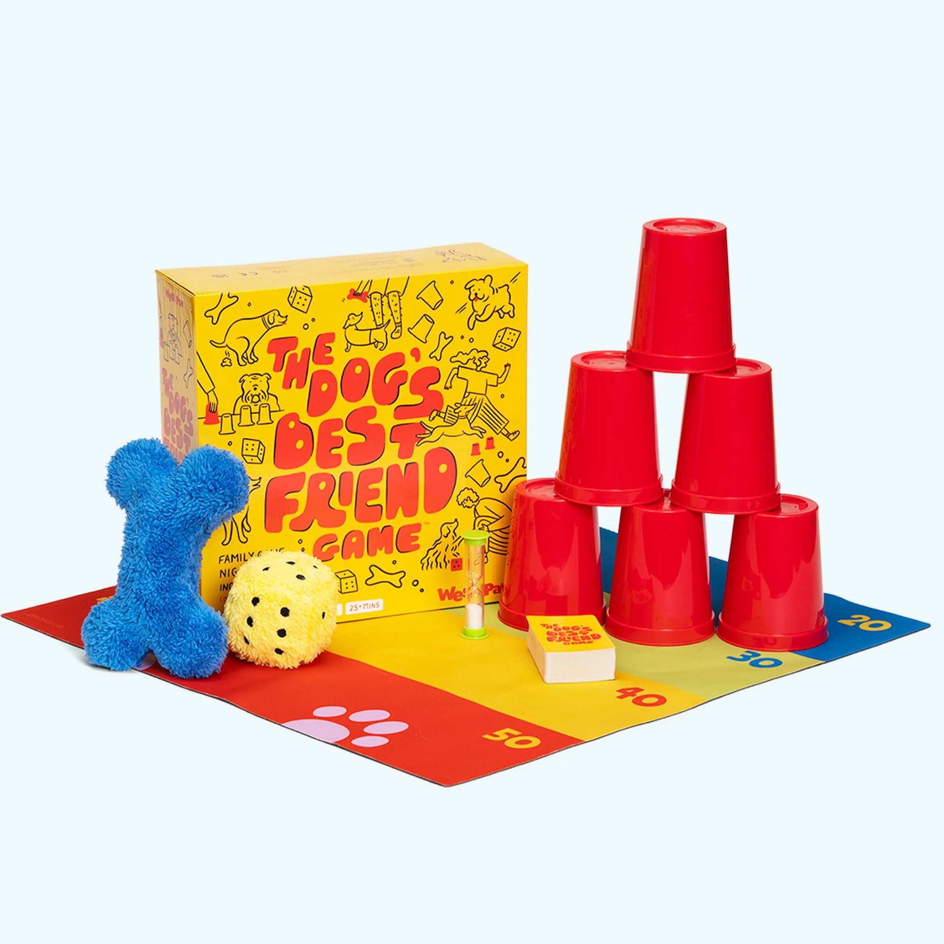 "The Dog's Best Friend" board game box with a blue plush bone toy, a yellow plush dice toy, 6 red cups stacked in a tower shape, a sand timer, playing cards, all on a play mat